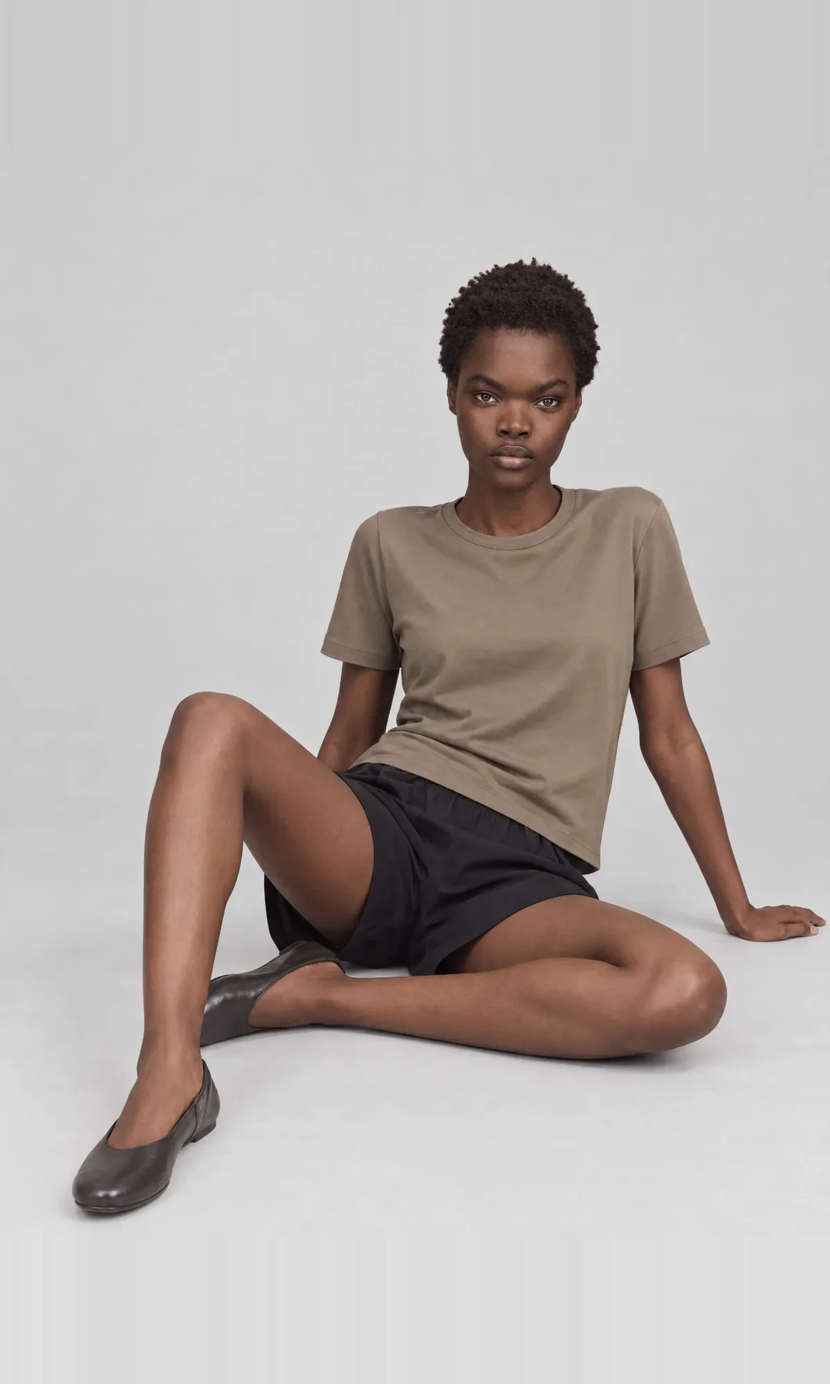 Seated catalogue view of a model wearing a taupe T-shirt and black shorts